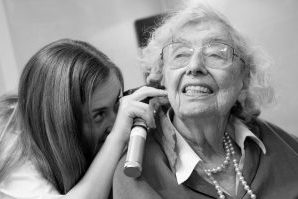 An elderly woman is smiling while she has her right ear examined by a student clinician
